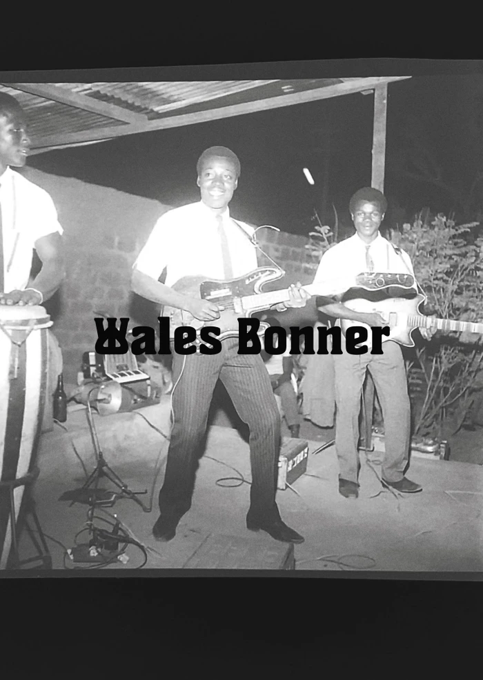Black and white image of three men playing electric guitars and drums outdoors at night under a covered structure.