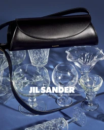 A sleek black leather handbag suspended above an arrangement of clear crystal glassware against a blue background.