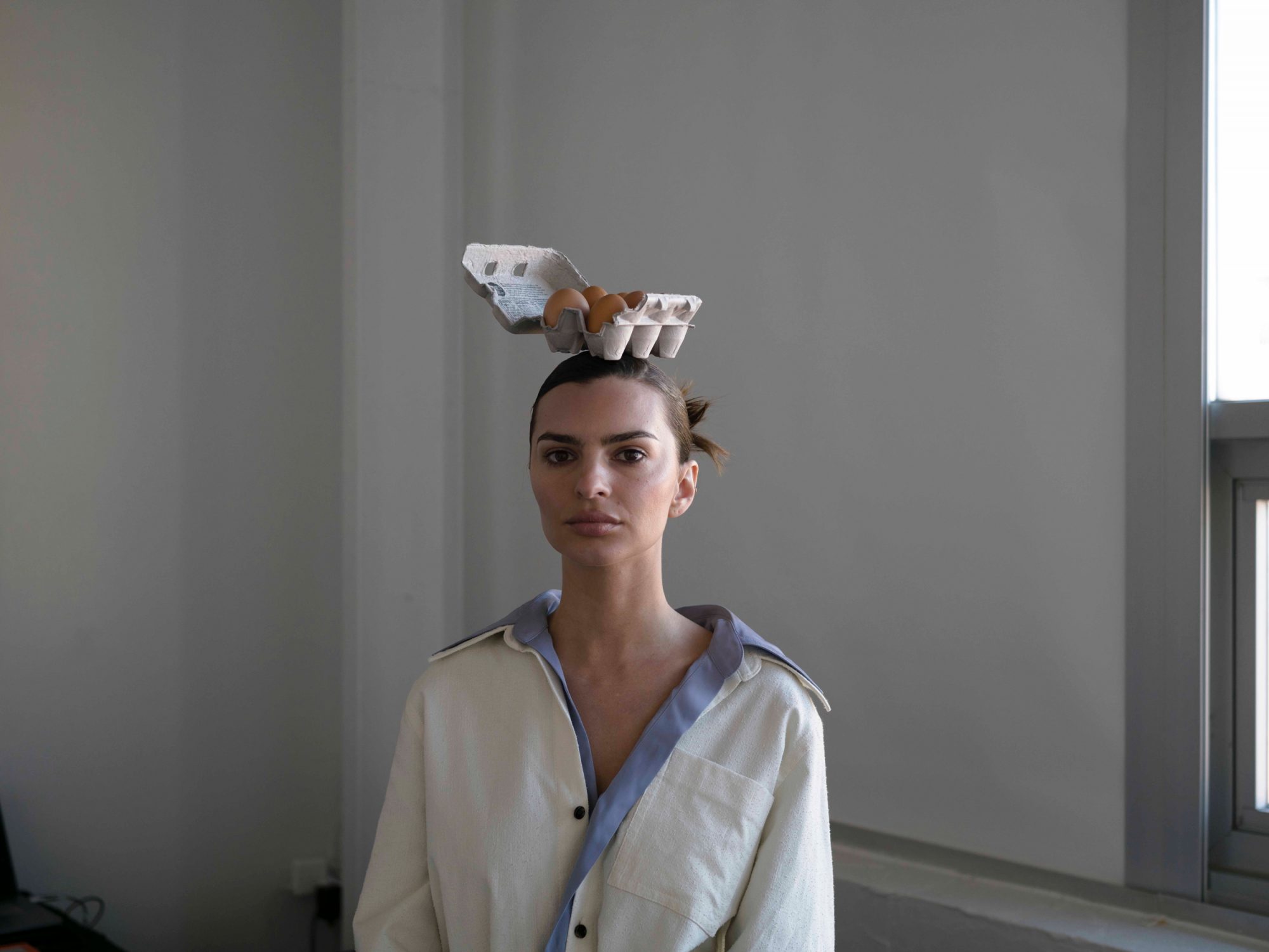 Portrait of a woman with an egg carton on her head, posing indoors in front of a window with neutral light, wearing a collared shirt.