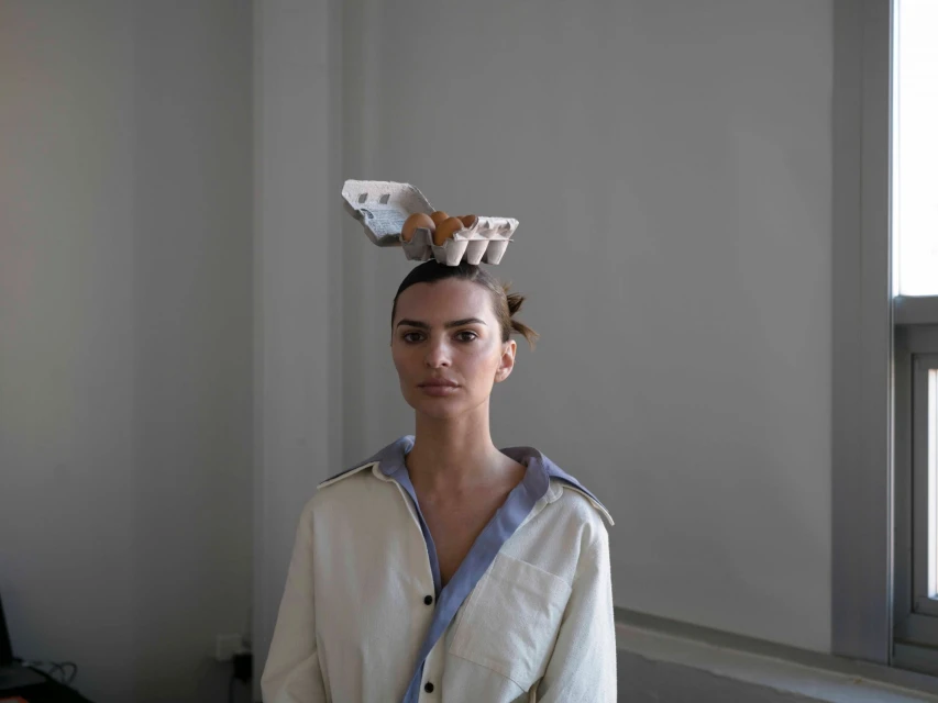 Portrait of a woman with an egg carton on her head, posing indoors in front of a window with neutral light, wearing a collared shirt.