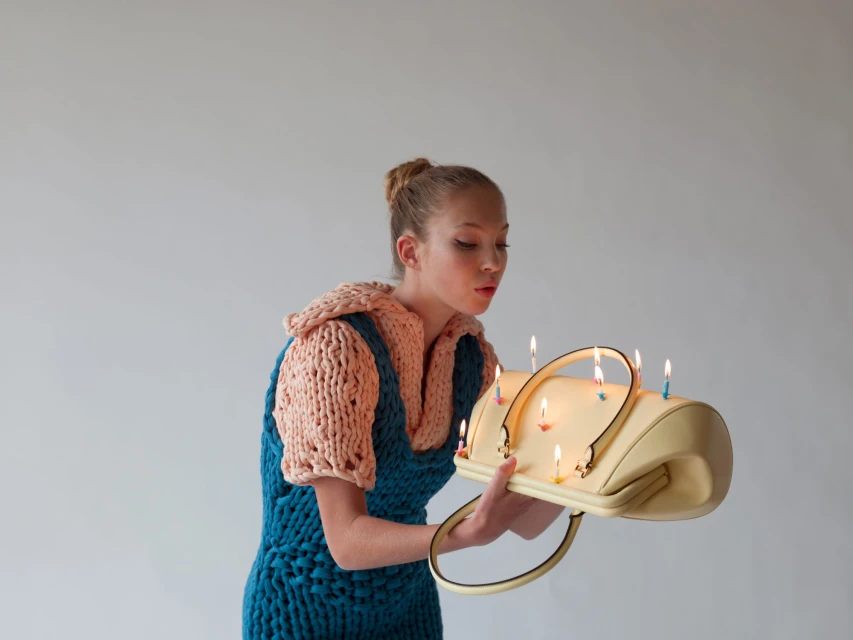 A fair-skinned young woman with a bun blows out candles in a yellow handbag, wearing an oversized knit outfit in a studio setting.