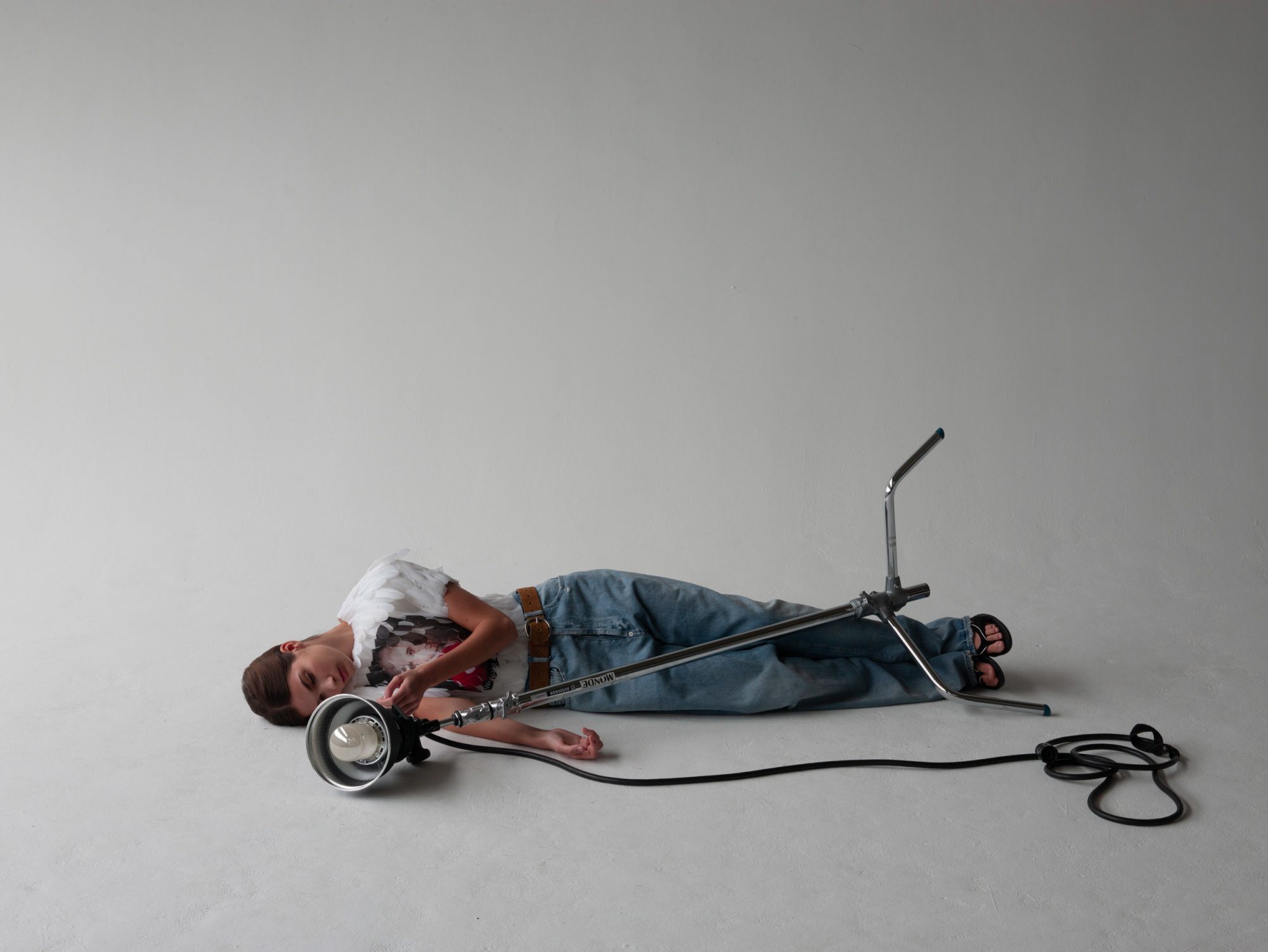 Young boy lying down next to studio lighting equipment in a light gray photo studio with a power cord, wearing jeans and a graphic tee.