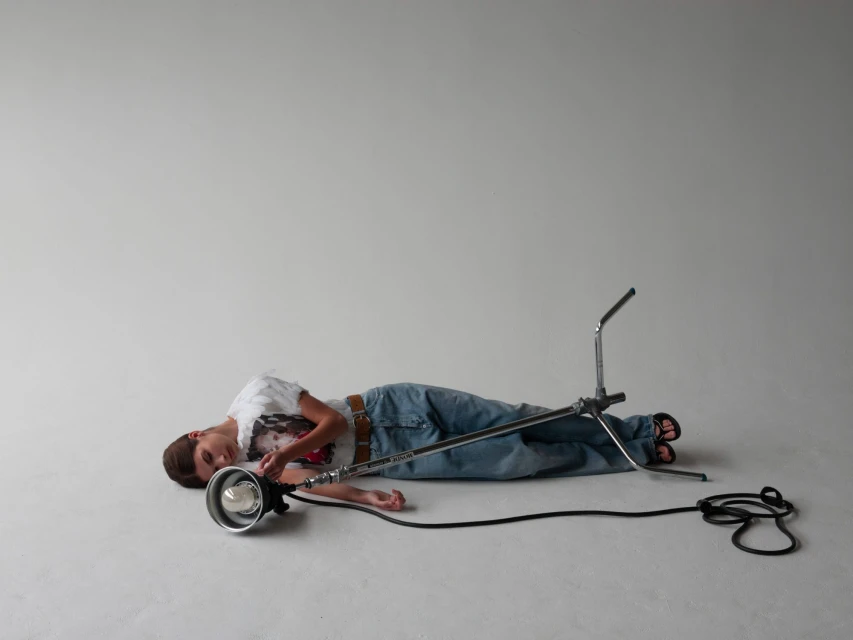 Young boy lying down next to studio lighting equipment in a light gray photo studio with a power cord, wearing jeans and a graphic tee.