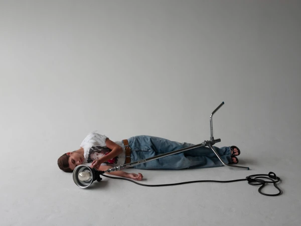 Young boy lying down next to studio lighting equipment in a light gray photo studio with a power cord, wearing jeans and a graphic tee.