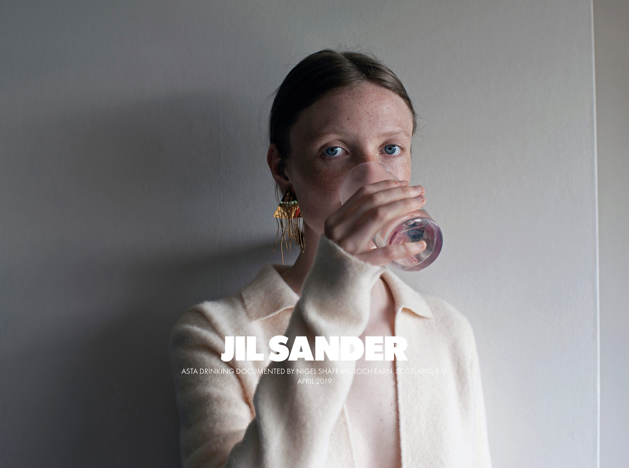 Woman drinking from glass, wearing a cream-colored cardigan and gold dangle earrings, against a neutral wall. Fashion photography.