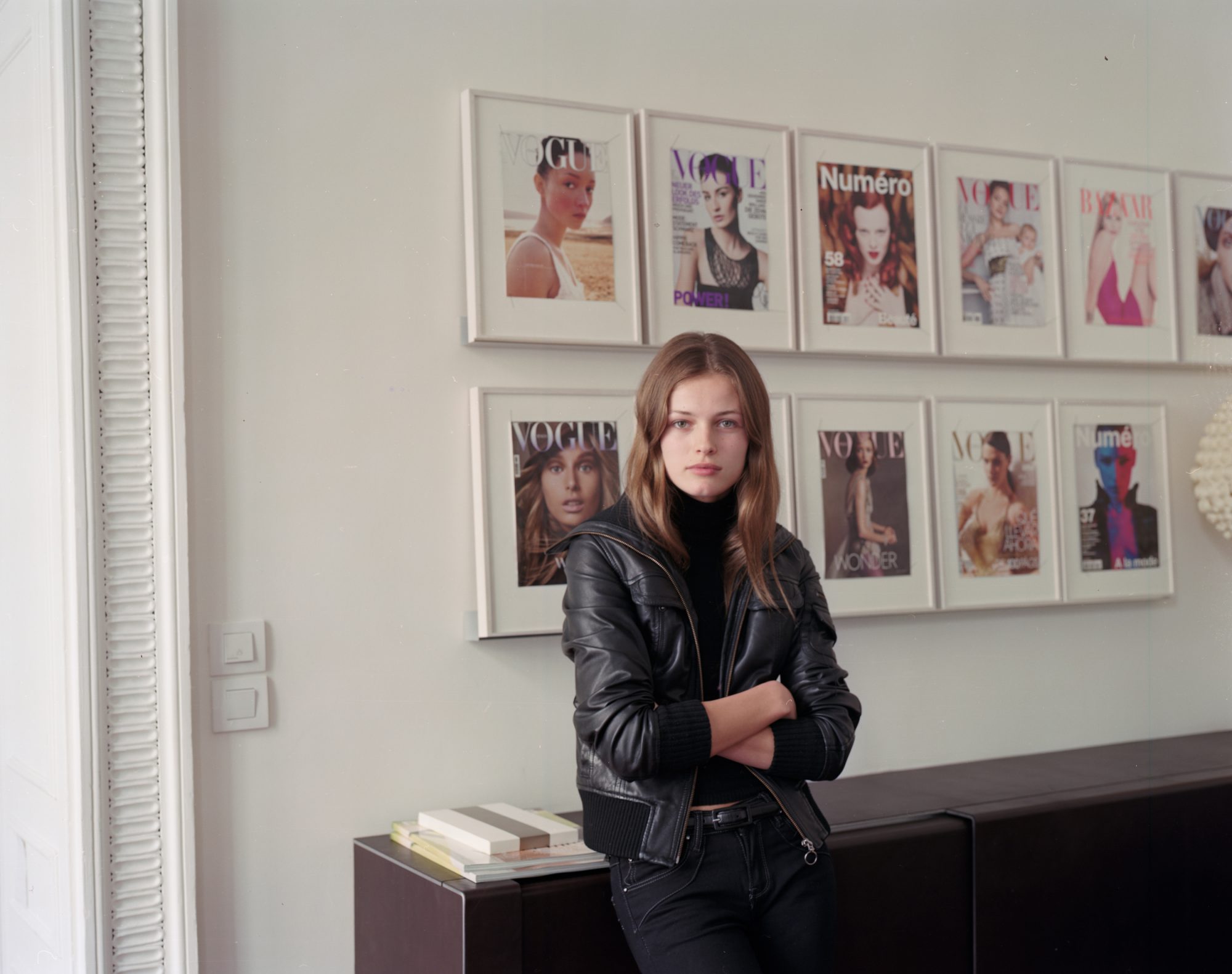 Young woman in a leather jacket stands in front of magazine covers, creating a fashion-forward and editorial-style composition indoors.
