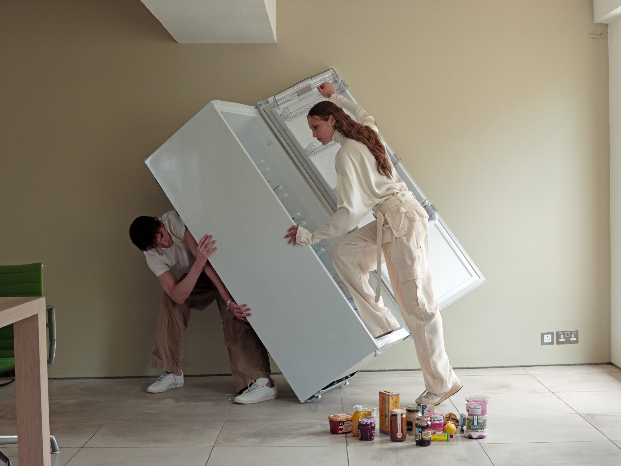 Two people moving a white refrigerator with food jars and containers on the floor inside a room with beige walls and tile floor.