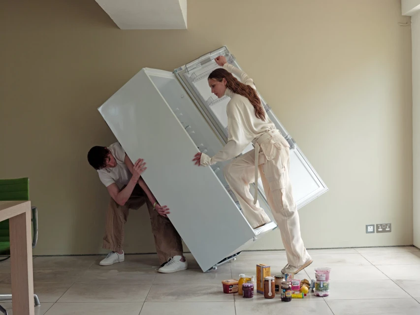 Two people moving a white refrigerator with food jars and containers on the floor inside a room with beige walls and tile floor.