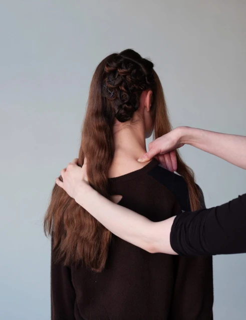 Woman with brown hair being styled with a braid. A second person is touching her shoulder and neck in a light studio setting.