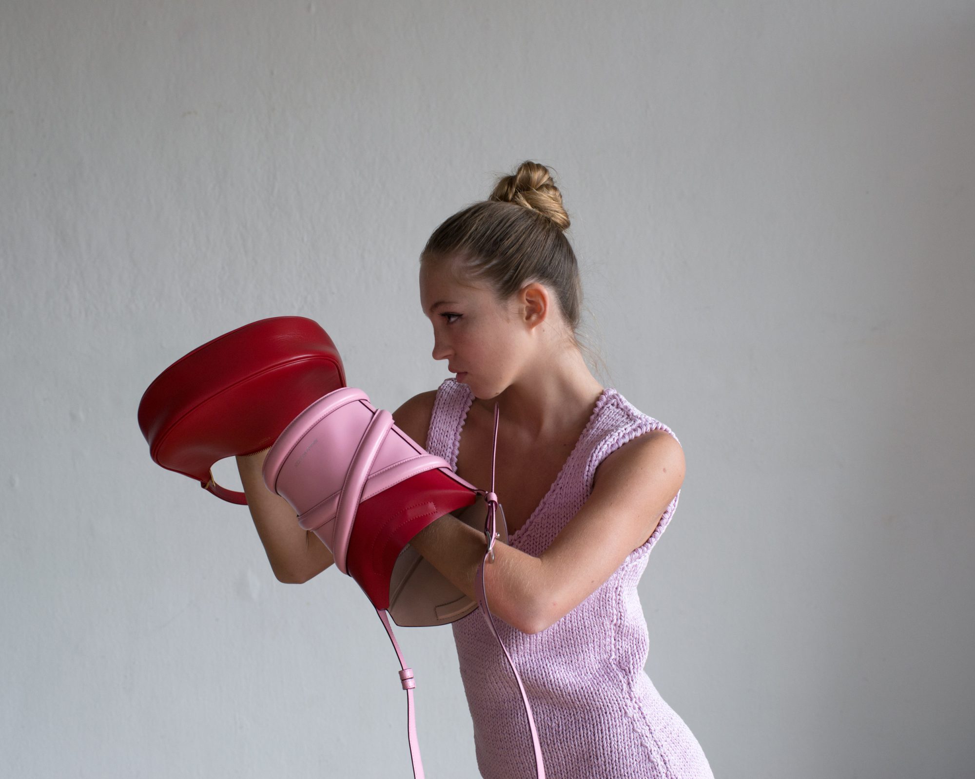 Young woman in knitted dress poses in studio with two red and pink designer handbags in front of her face, fashion forward look.