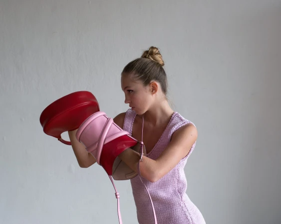 Young woman in knitted dress poses in studio with two red and pink designer handbags in front of her face, fashion forward look.