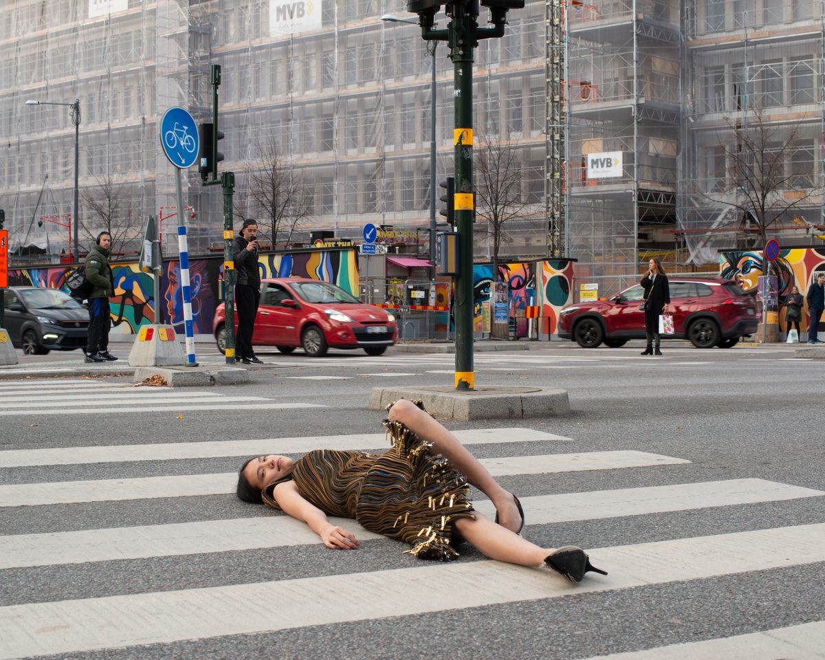 Adult female in dress lies prone on zebra crossing in urban setting with cars and pedestrians in background, overcast daylight.