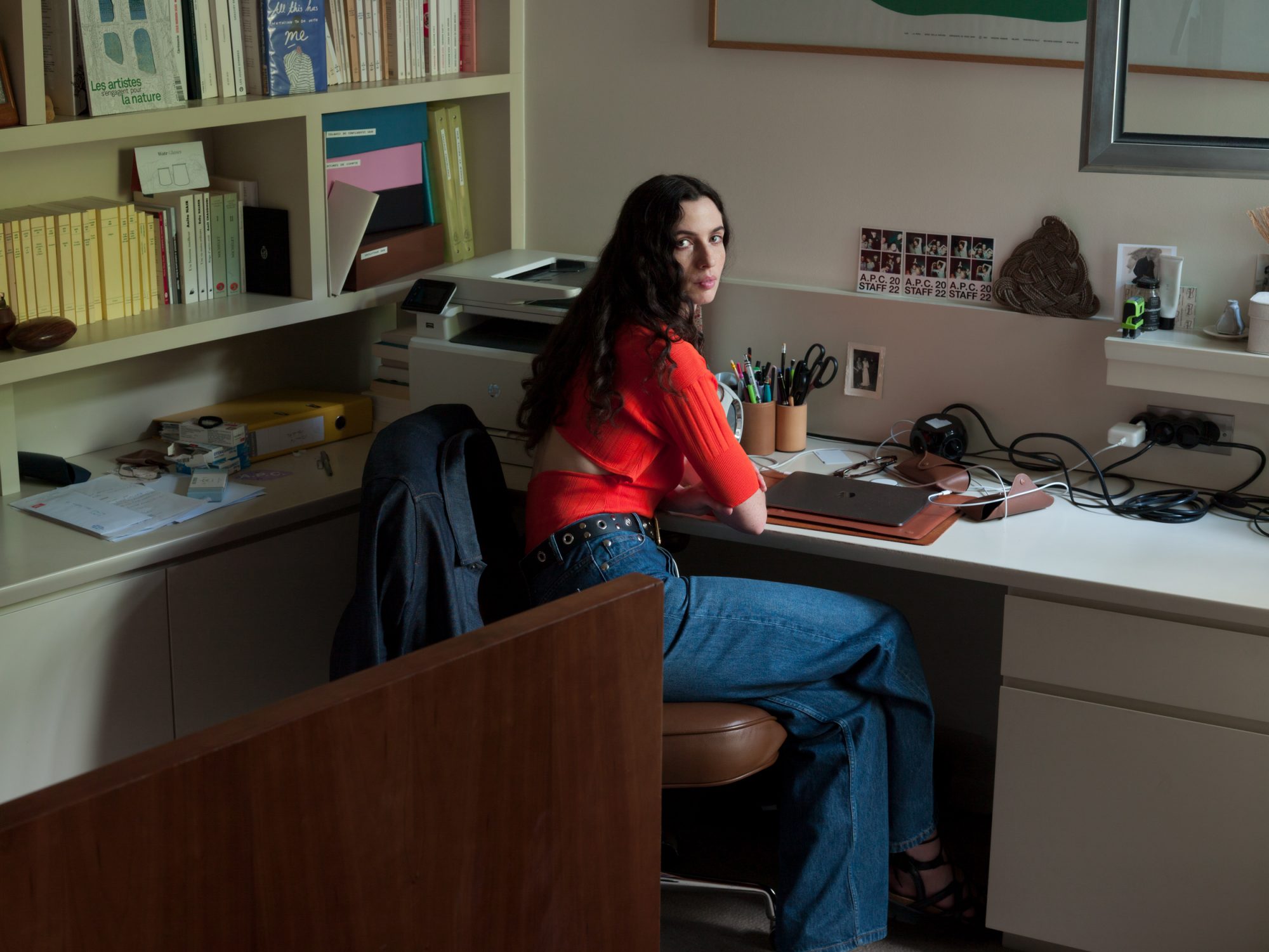 Young woman with long dark hair sits at a desk with a laptop in an office setting, illuminated by daylight and desk lighting.