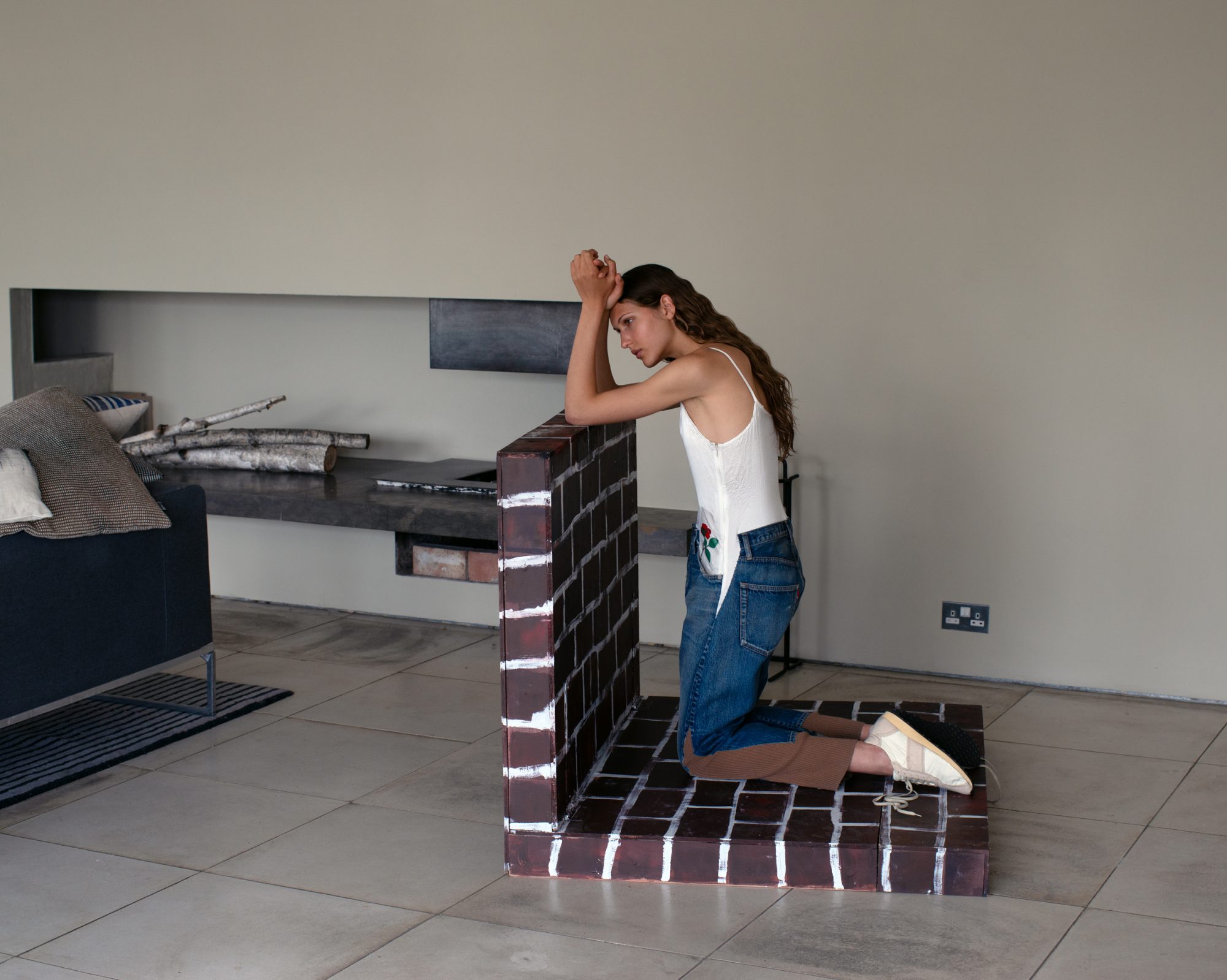 A woman in a white top and blue jeans kneels next to a faux brick structure in a neutral-toned interior, featuring a modern fireplace detail and minimal decor.