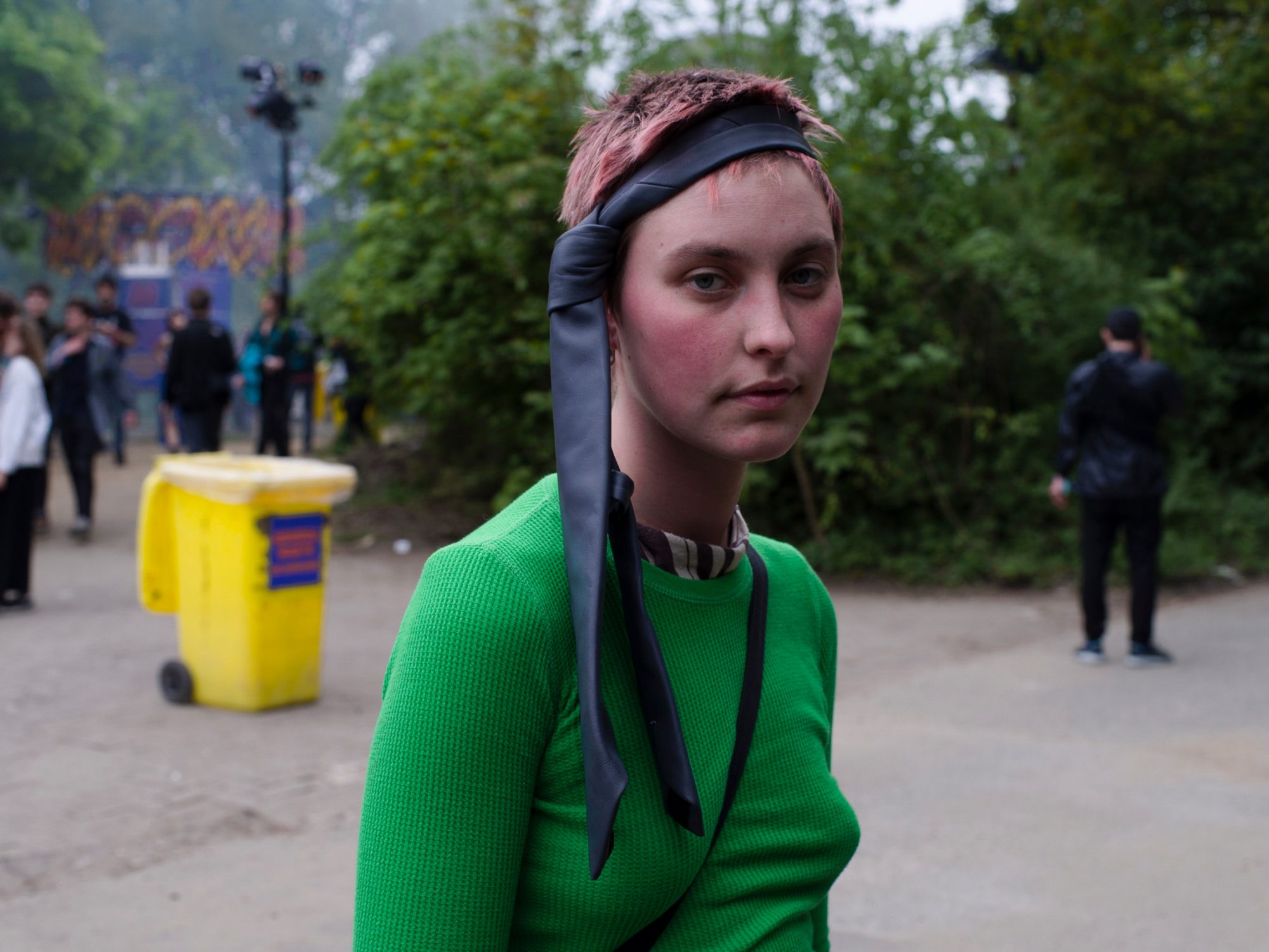 Portrait of a young adult female with short pink hair, wearing a green top and black headscarf, standing outdoors with a blurred background.