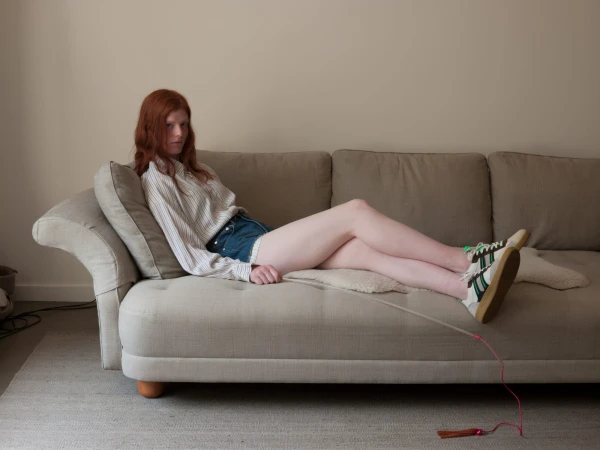 Young woman with red hair, wearing denim shorts and a striped shirt, relaxes on a light beige couch in an indoor living space.