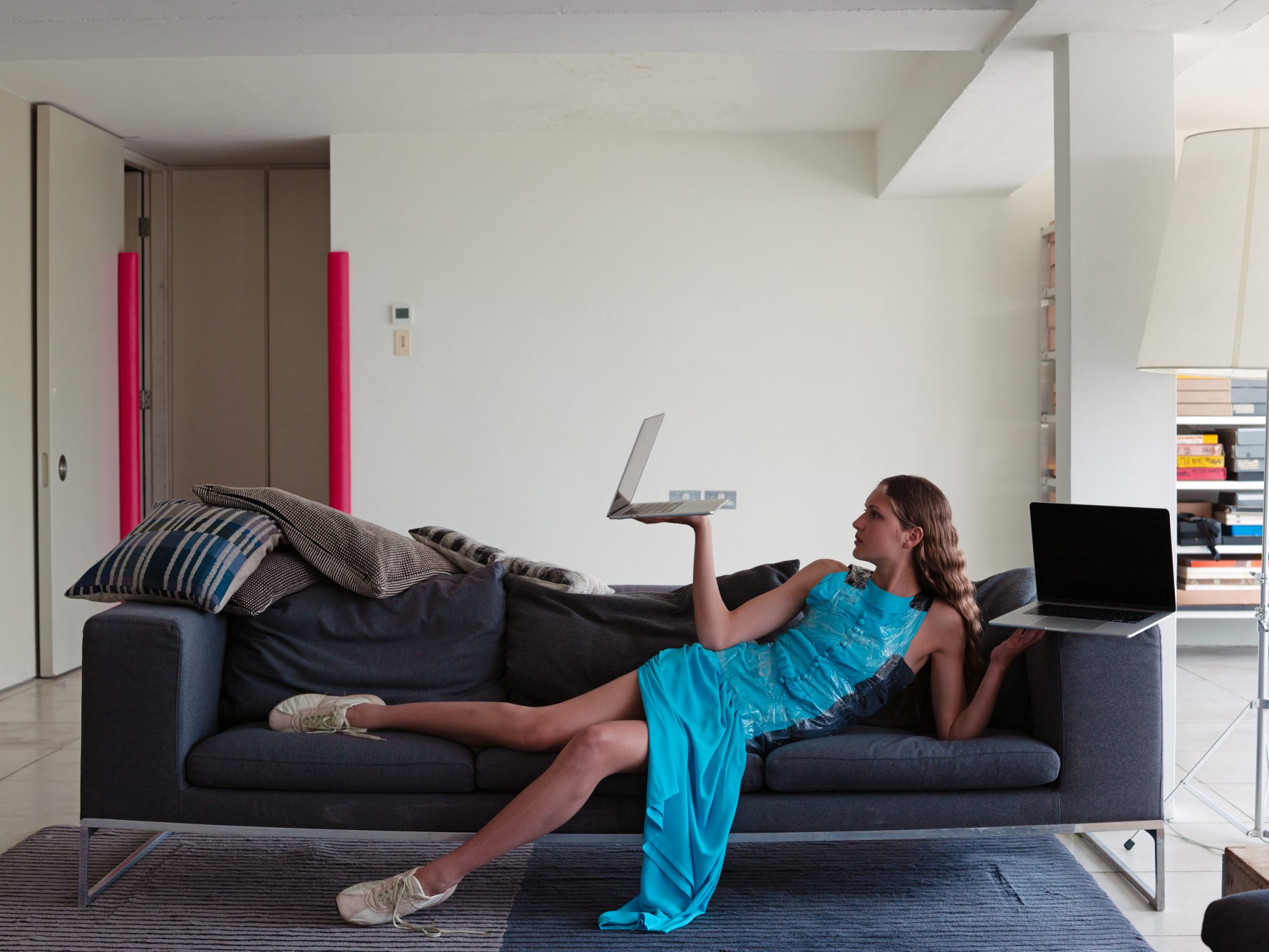 A woman lying on a grey couch wearing a blue dress and white sneakers holds two laptops in a brightly lit interior living space.
