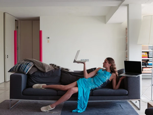 A woman lying on a grey couch wearing a blue dress and white sneakers holds two laptops in a brightly lit interior living space.
