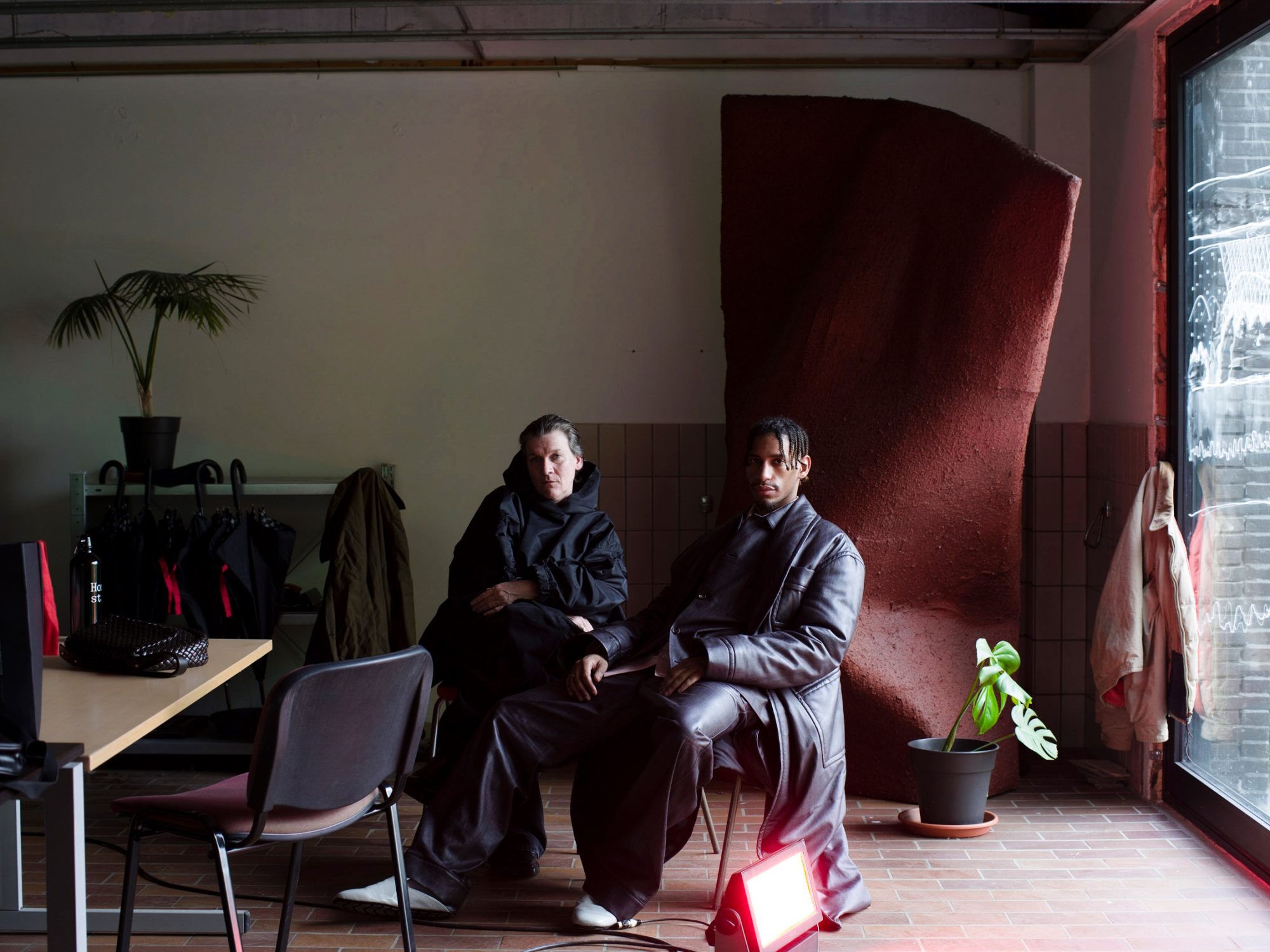 Two people sit indoors in an industrial-style space near a sculpture, plant, and window, lit by a red light, showcasing casual fashion.