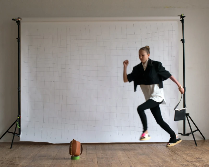 A young adult female fashion model is frozen mid-air in a studio, running and holding a purse, with a vintage bag on the floor.