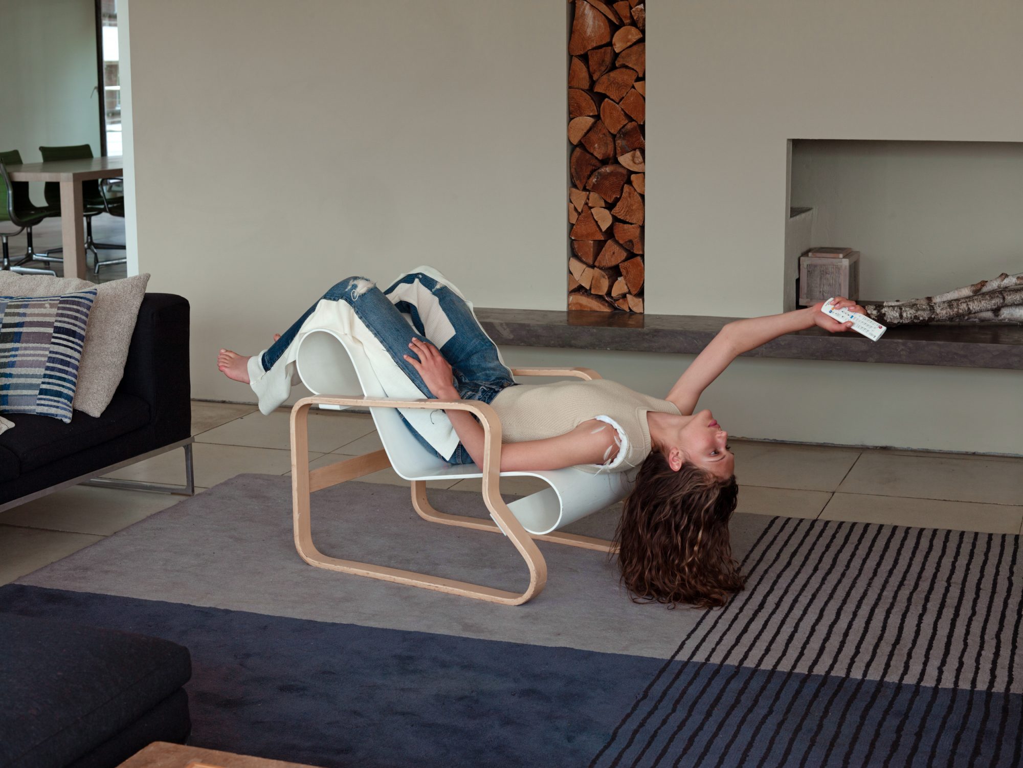 Woman lounges upside down on modern chair in bright living room with remote control. Wood and birch textures, neutral colors.