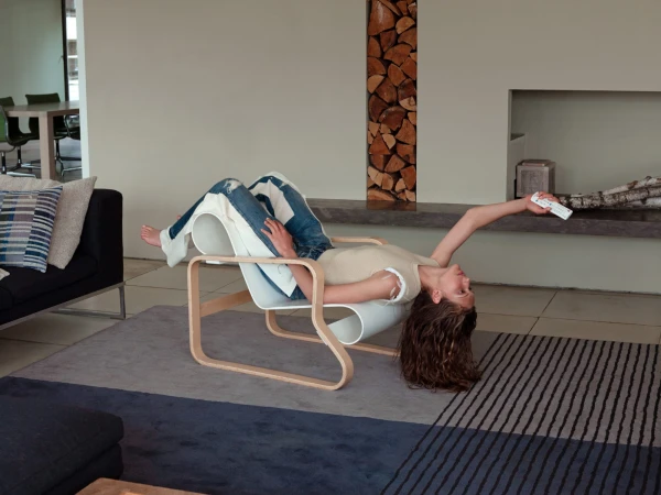 Woman lounges upside down on modern chair in bright living room with remote control. Wood and birch textures, neutral colors.