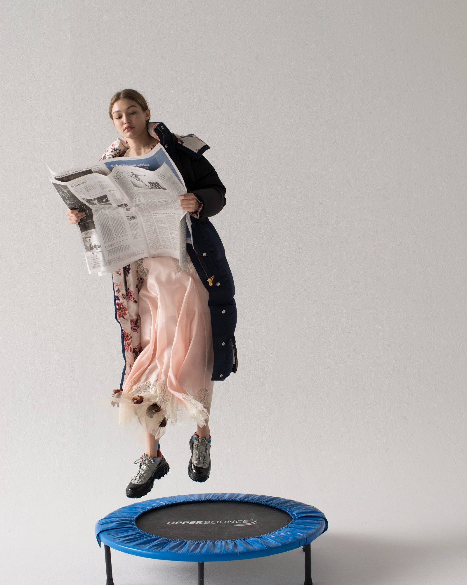Blonde-haired woman in a floral dress and puffer jacket floats above a trampoline reading a newspaper in a bright studio setting.