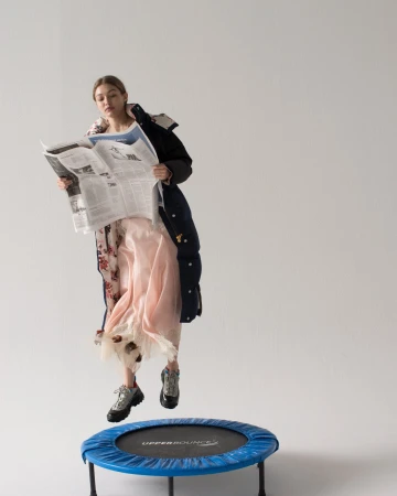 Blonde-haired woman in a floral dress and puffer jacket floats above a trampoline reading a newspaper in a bright studio setting.
