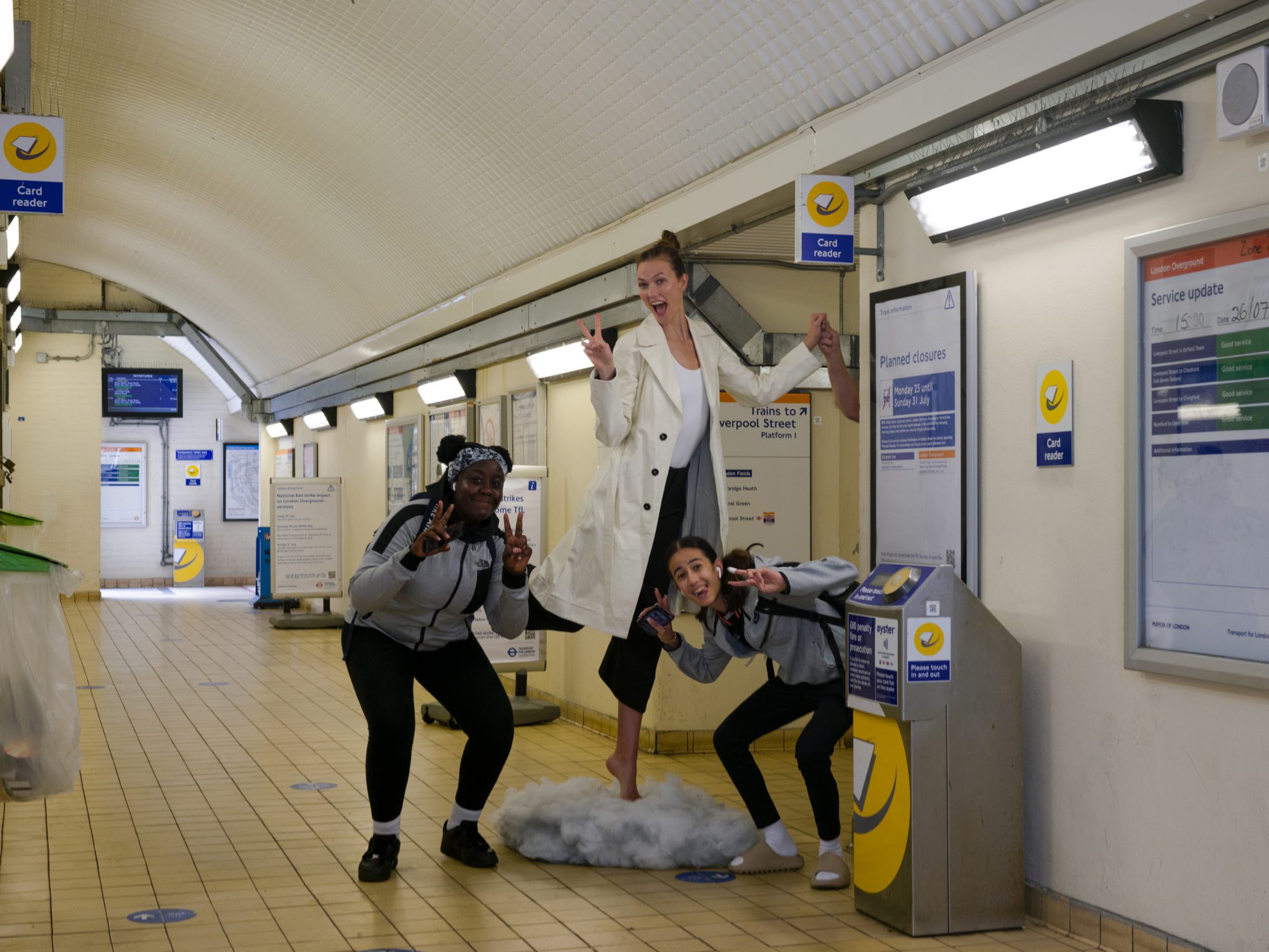 Three people pose playfully in a train station, one standing on a faux cloud, showcasing a blend of candid fun and urban transit scenery.