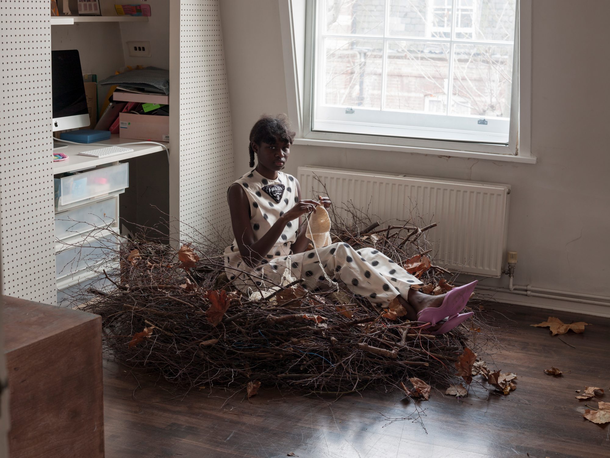 Fashionable woman poses in a nest of branches in a room with computer desk and radiator near window providing natural light.