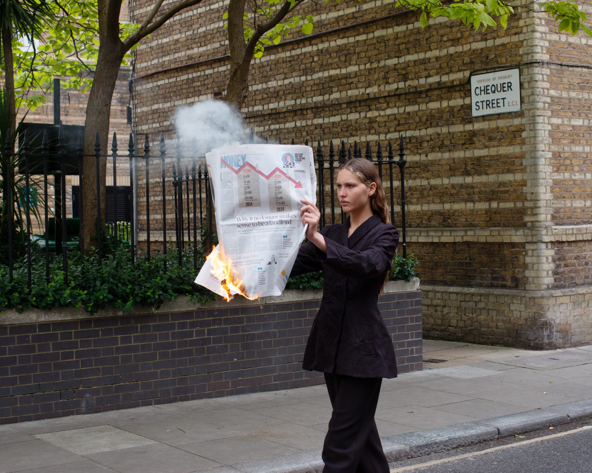 Woman in a suit holds a burning newspaper on a city street. The newspaper has a graph on it and smoke is rising.