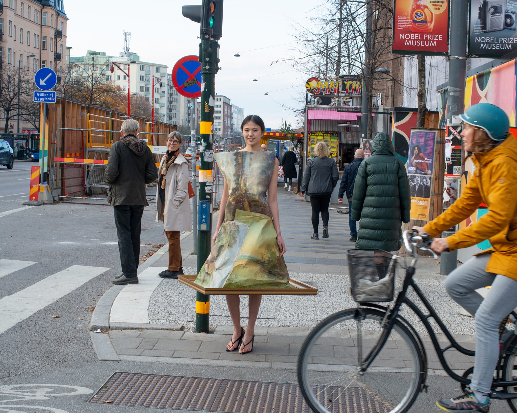 Young woman wearing an artful paper dress standing on a sidewalk with passersby and a cyclist in an urban environment during daytime.