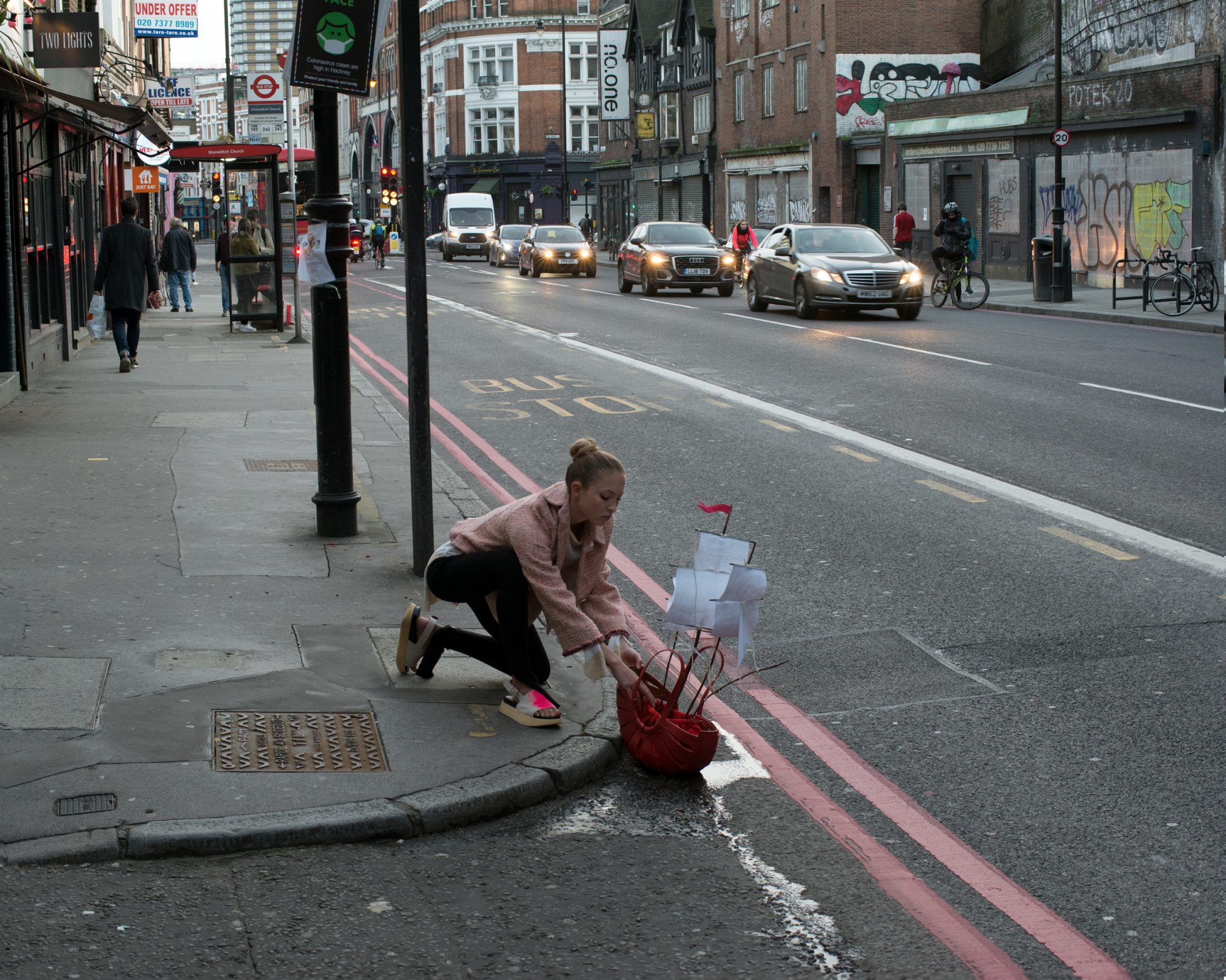Woman crouches near a toy ship on the side of a busy urban street in London, with cars, buildings, sidewalks and pedestrians in view.