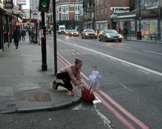 Woman crouches near a toy ship on the side of a busy urban street in London, with cars, buildings, sidewalks and pedestrians in view.