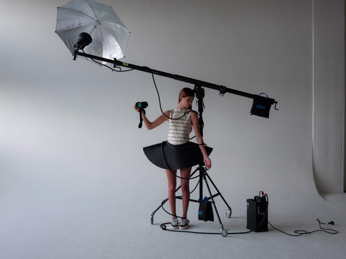 Young woman in whimsical black-and-white dress holds a camera, standing near lighting equipment in a bright photo studio.