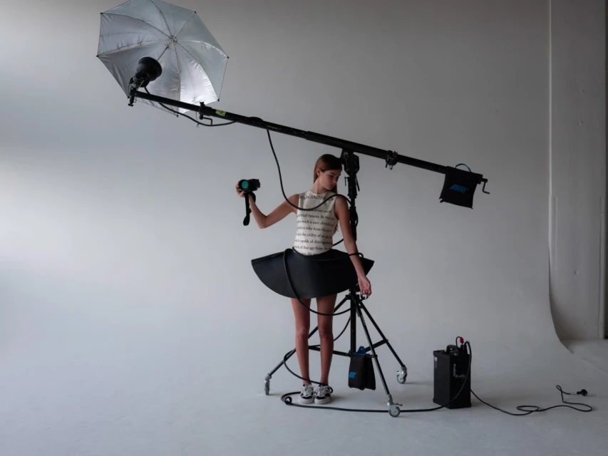 Young woman in whimsical black-and-white dress holds a camera, standing near lighting equipment in a bright photo studio.