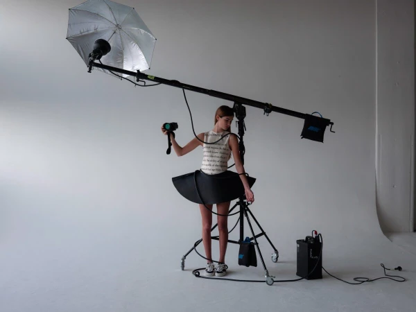 Young woman in whimsical black-and-white dress holds a camera, standing near lighting equipment in a bright photo studio.