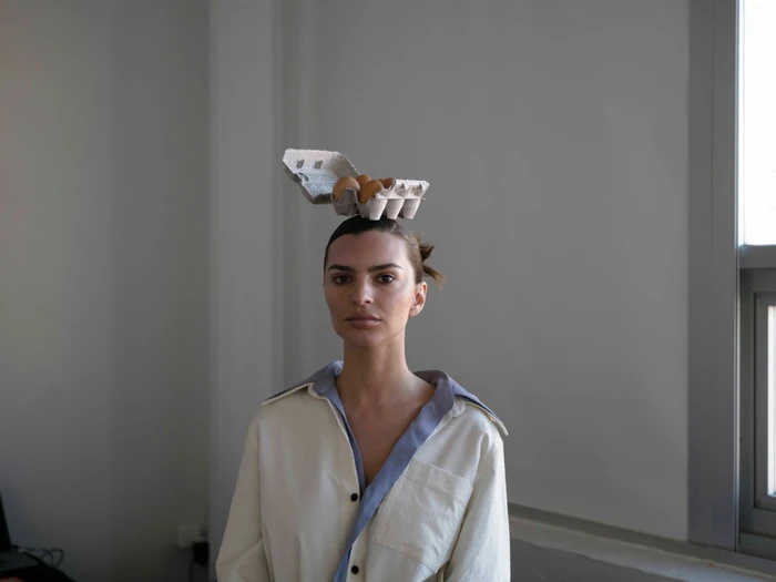 A woman balances an open carton of eggs on her head, wearing an off-white button-up shirt in an indoor setting.