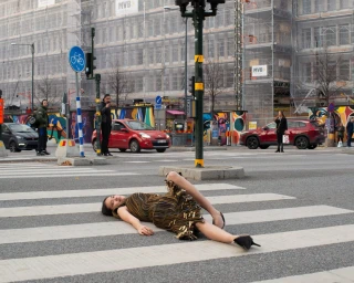 Woman wearing a patterned fringe dress lying on a pedestrian crosswalk on a city street.