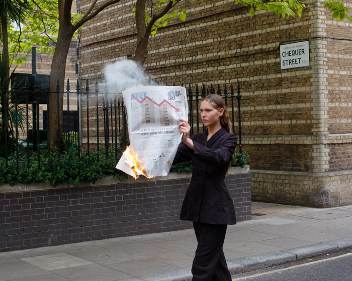 A woman in a black blazer holding a burning newspaper on a London street near a Chequer Street sign