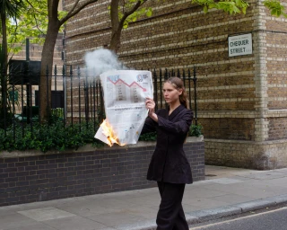 A woman in a black blazer holding a burning newspaper on a London street near a Chequer Street sign