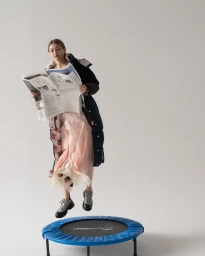 Model reading a newspaper while jumping on a small trampoline in a studio with a white backdrop.