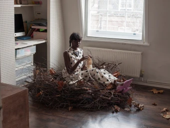 Black woman wearing polka-dot clothing sitting inside a large nest made of twigs and dry leaves in a room.