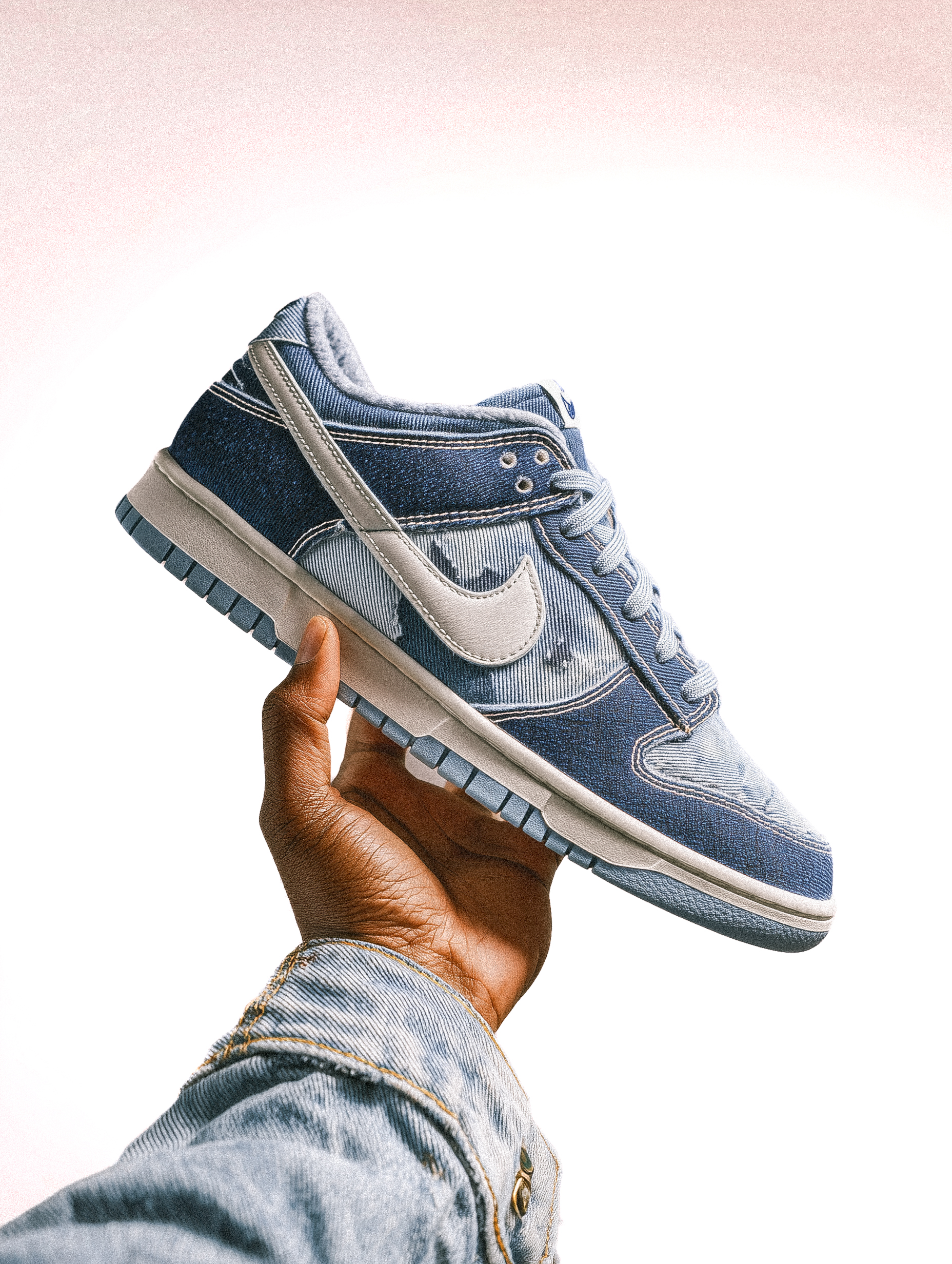 Denim Nike Dunk sneaker held in hand. Textured blue and white shoe in close-up with a denim-clad arm against a bright background.