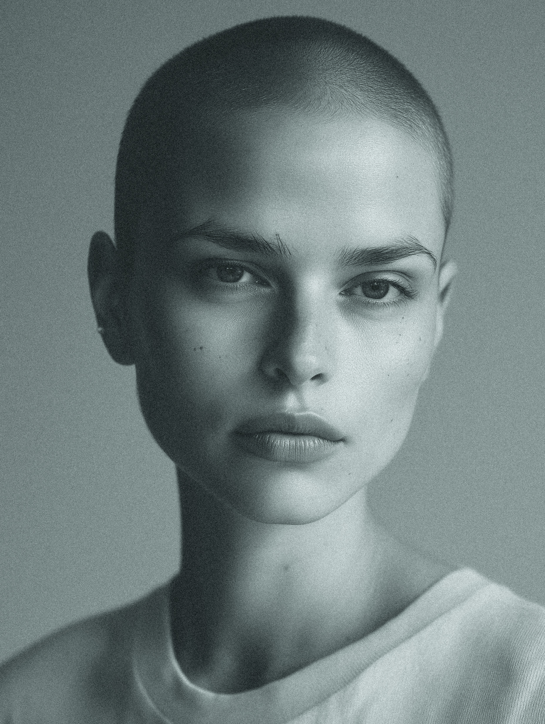 Close-up studio shot of a young adult woman with a shaved head, fair skin, and neutral expression wearing a light t-shirt against a grey background.