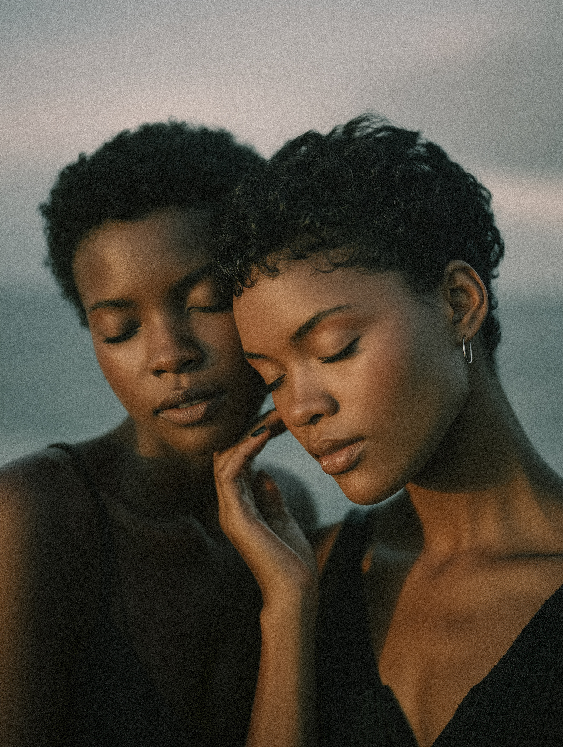 Close-up portrait of two dark-skinned women with short curly hair and closed eyes. They are wearing black tank tops against a diffused background.
