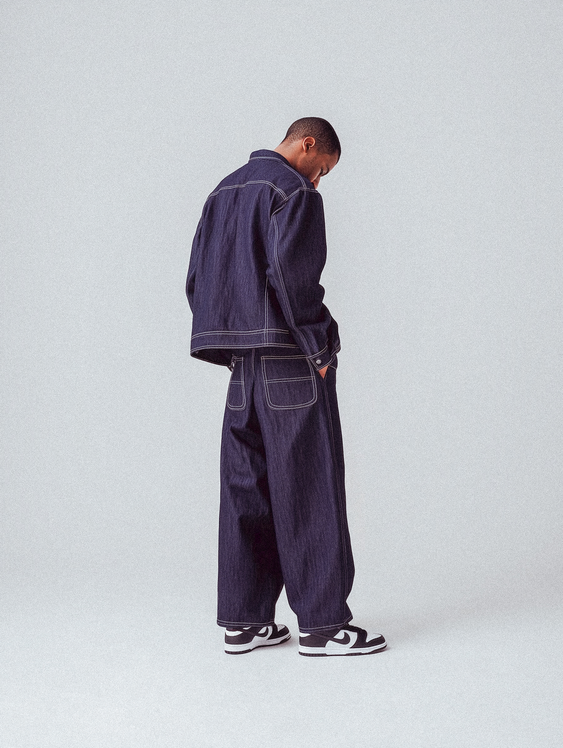 Denim fashion shoot featuring a slender adult male model in a matching denim jacket and baggy jeans set against a light gray background.