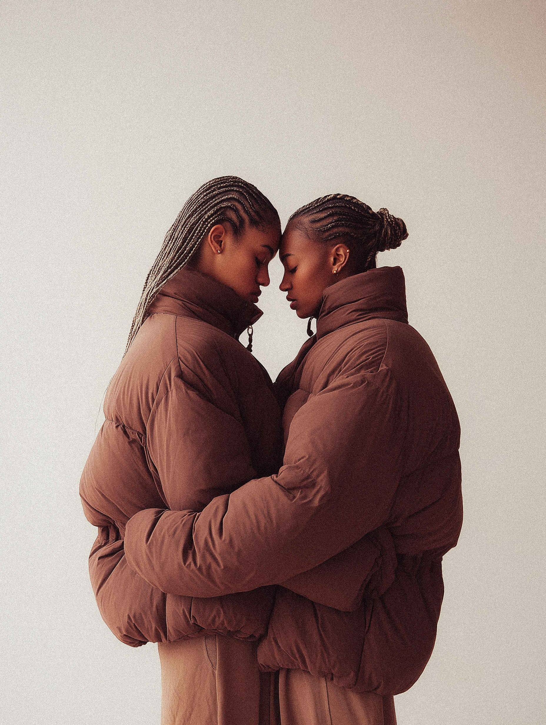 Two women in brown puffer jackets embrace in a close-up studio shot against a neutral background, showcasing fashion and connection.