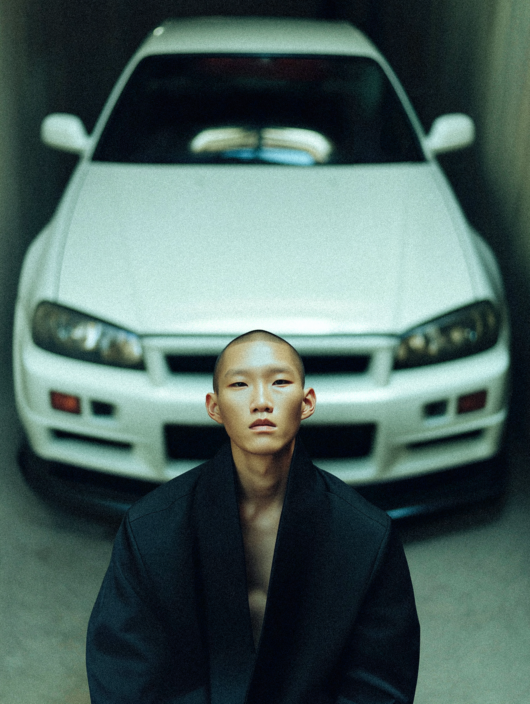 Man poses in black coat in front of a white Nissan Skyline R34 in an indoor garage setting with natural lighting.