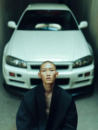 Man poses in black coat in front of a white Nissan Skyline R34 in an indoor garage setting with natural lighting.
